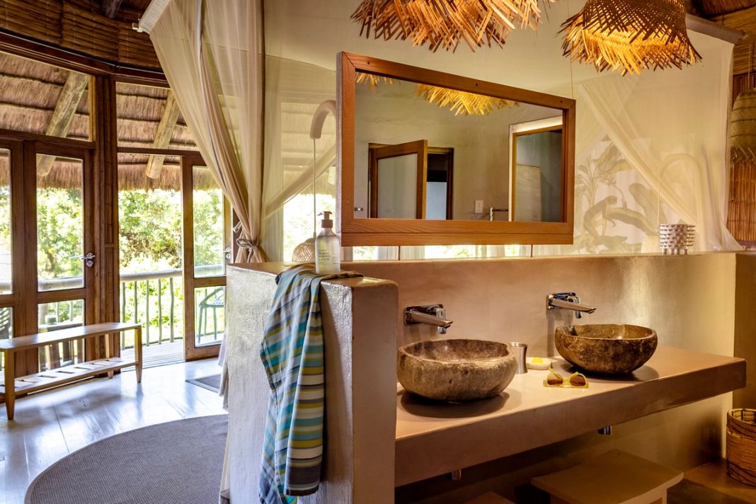 A forest-room bathroom with twin stone basins, a botanical print and glass doors onto a green-bush deck.