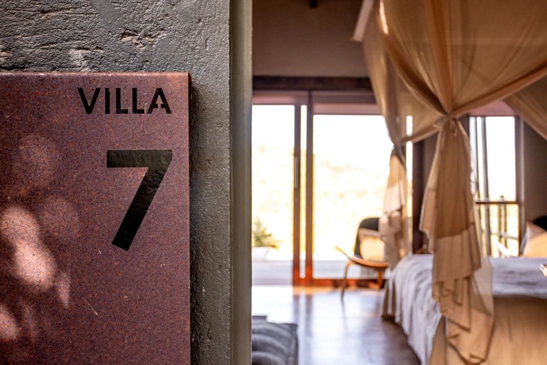 A rust-toned entrance wall reading Villa 7 beside a glimpse of a canopy-bed room and deck doors.