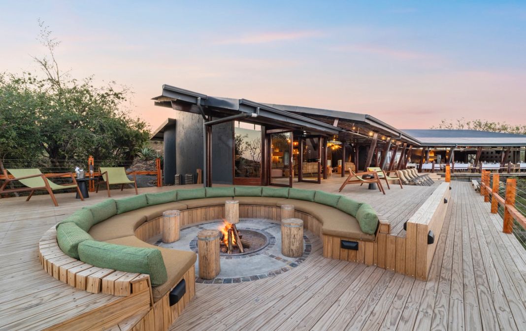 A circular fire pit with cushioned benches sits on the deck at dusk, with the lodge behind.