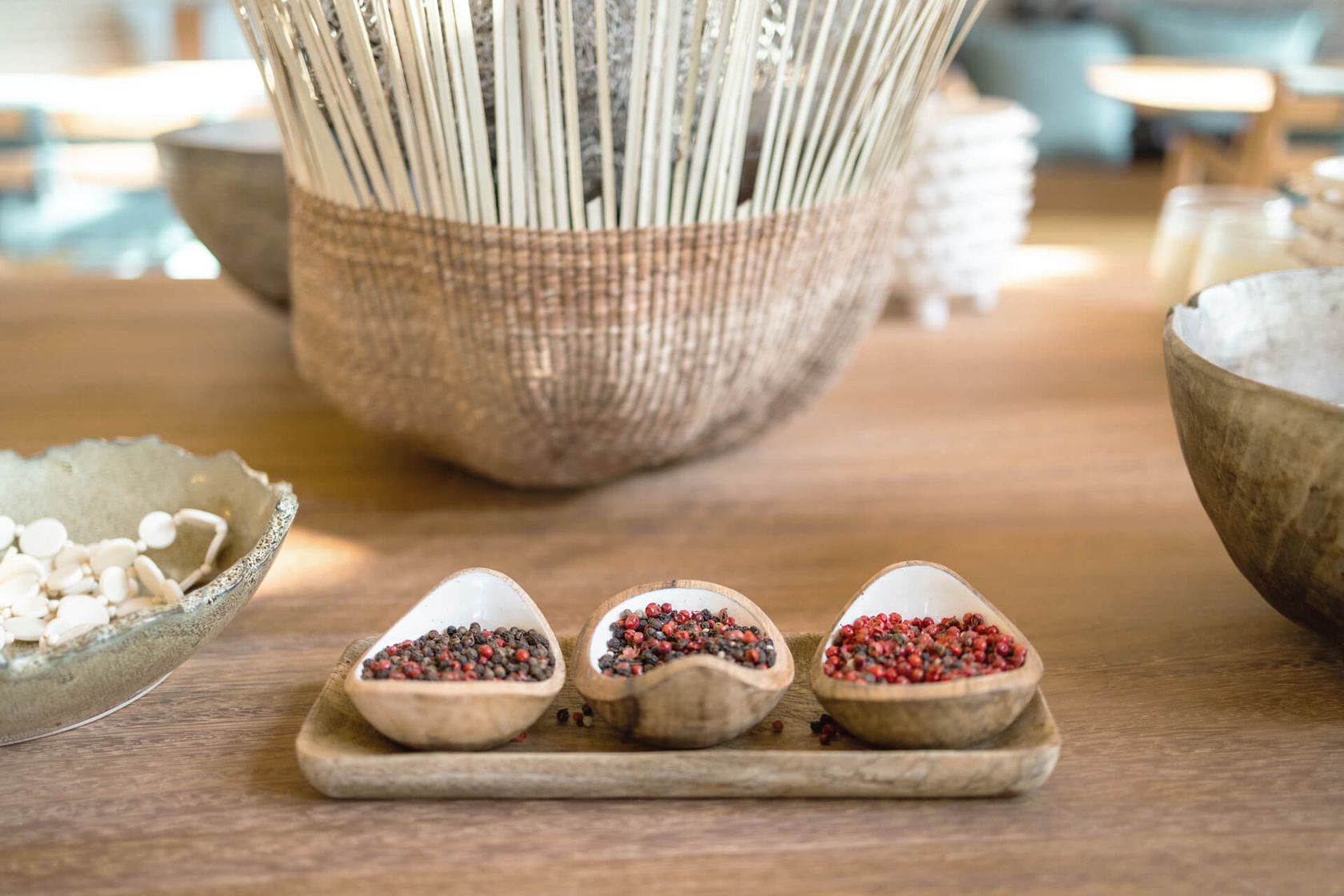 Three wooden pinch bowls of pink and black peppercorns on a tray, a woven basket and pale bowl behind.