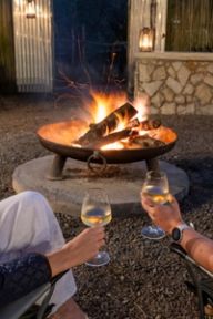 Two guests hold glasses of white wine beside the boma fire pit at dusk.