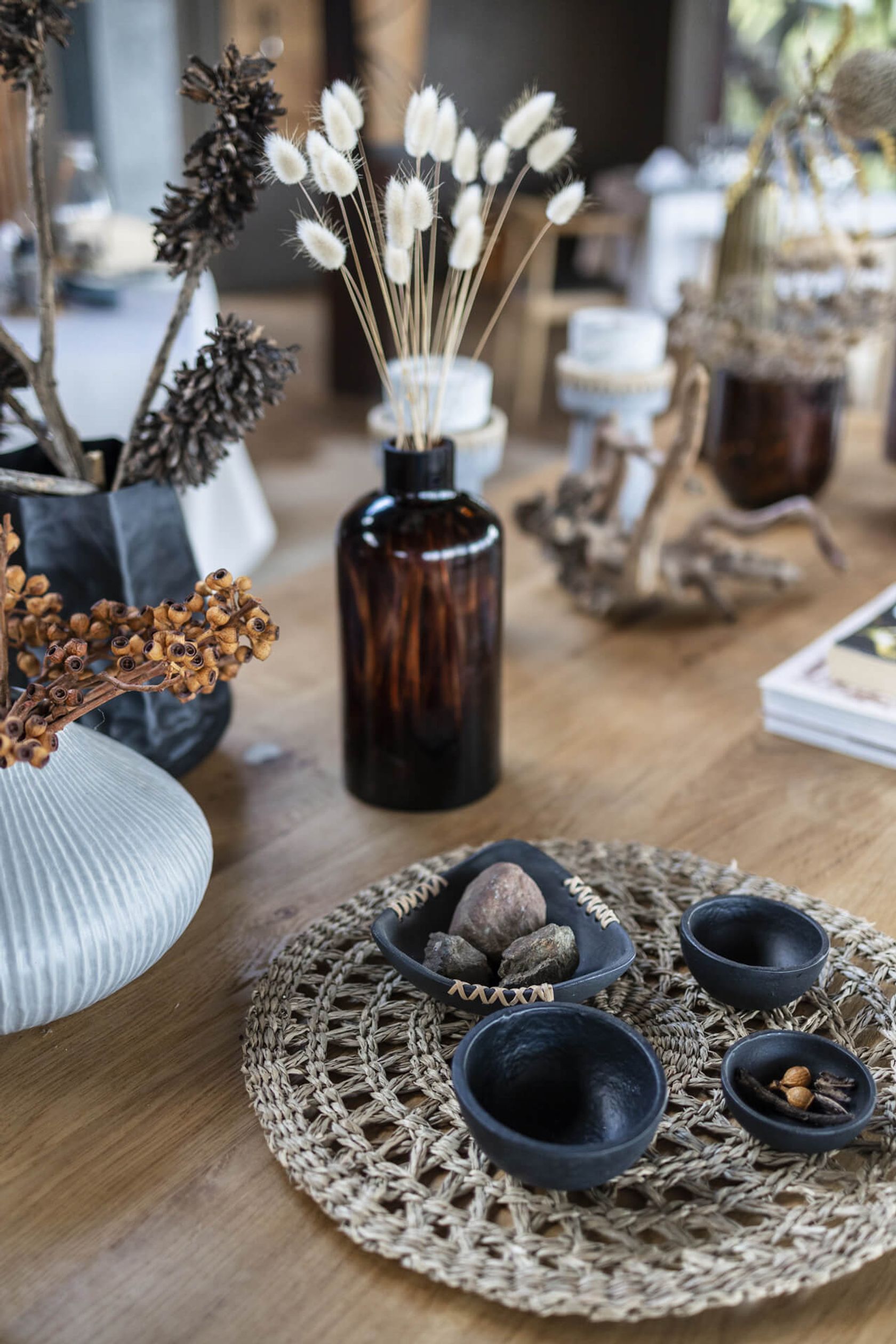 Dried grasses in an amber glass bottle stand beside dark stone bowls of spices on a woven table mat.