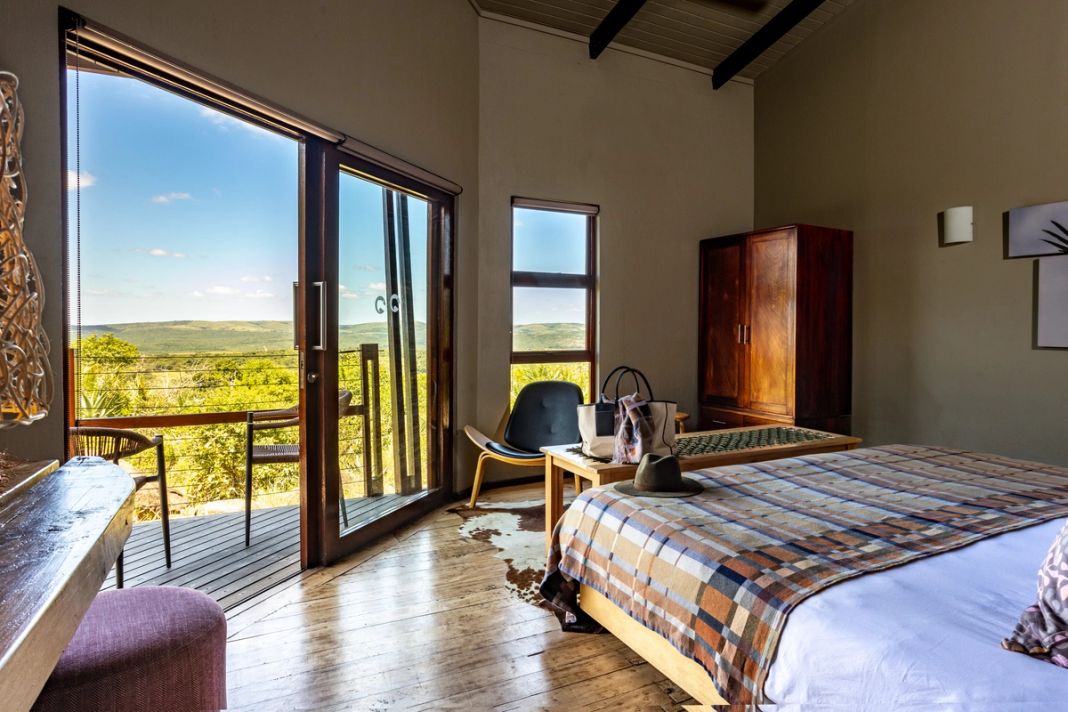 A safari room with a plaid-throw bed, a timber wardrobe and glass doors to a deck with a hill view.
