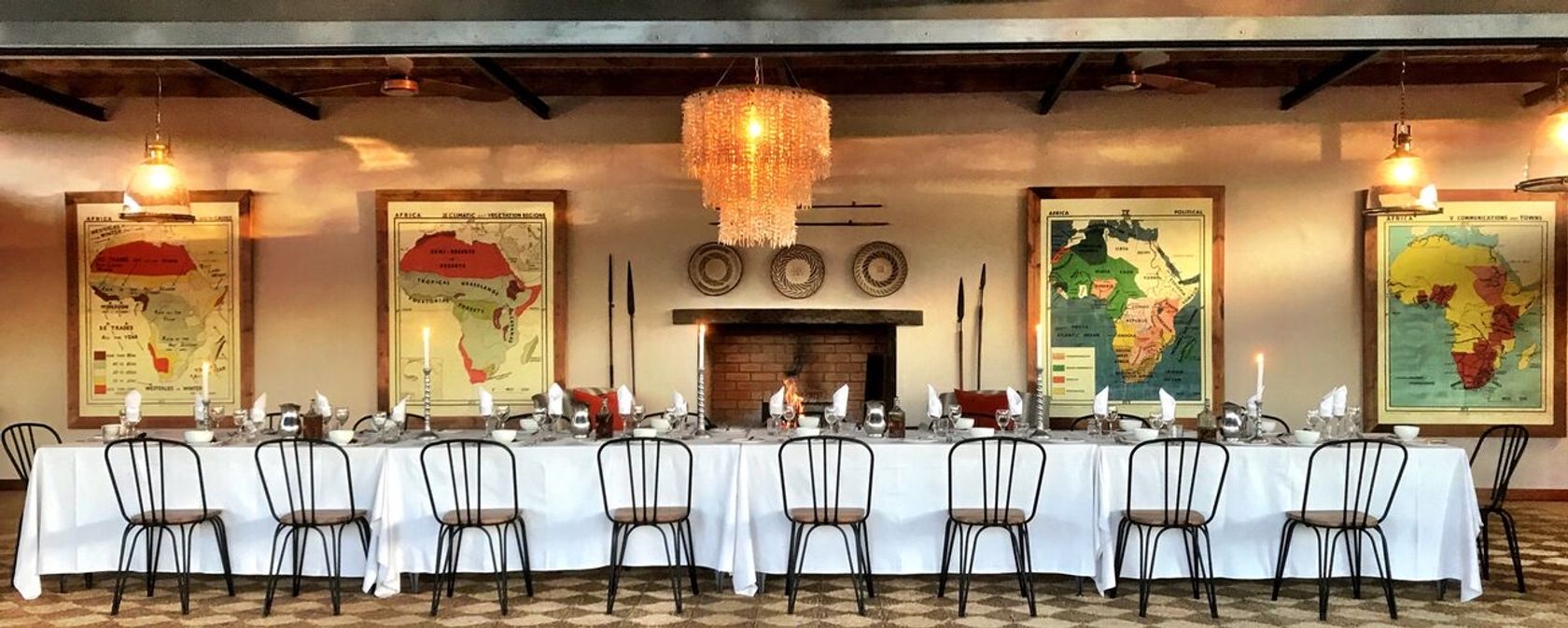 A long dining table set with white linen and candles below four vintage maps of Africa, a brick fireplace, and a beaded chandelier.