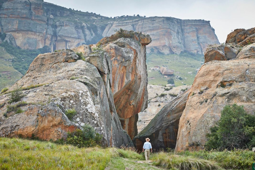 A lone hiker in a hat stands small at the foot of towering weathered sandstone cliffs in green veld.