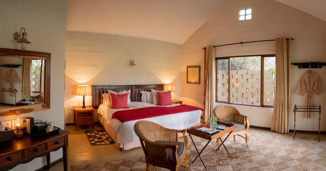 A guest house bedroom with a king bed under a vaulted ceiling, whitewashed brick walls, wicker chairs and a dressing table with a mirror.