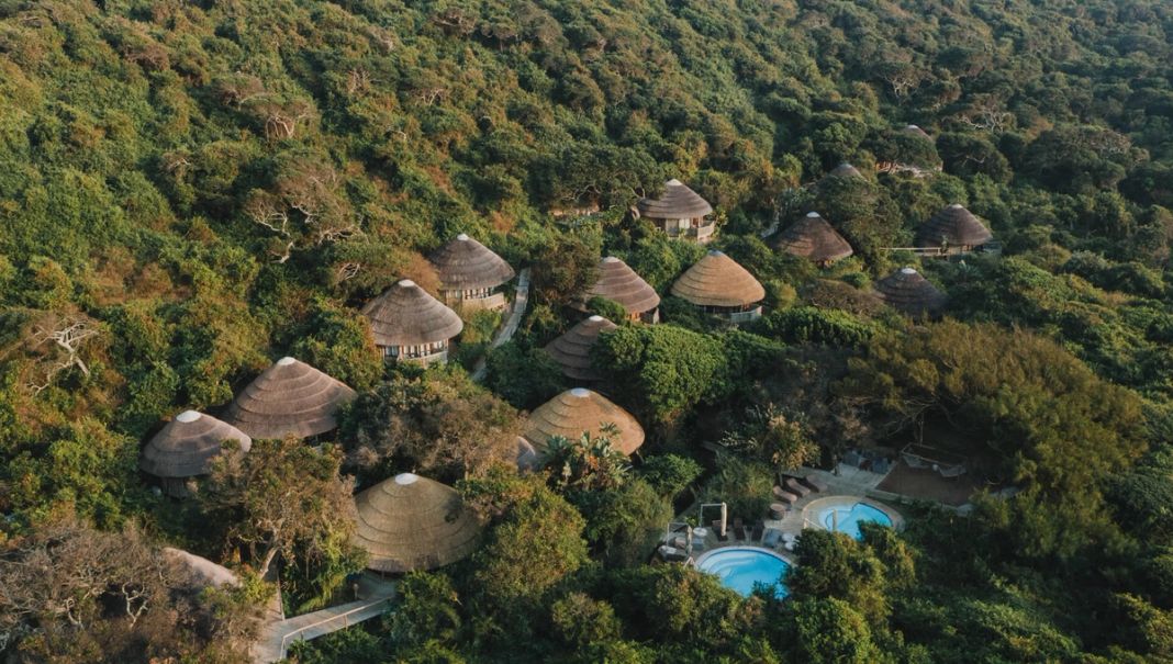 A golden-hour aerial of the lodge, a dozen conical thatched roofs in the dune forest above the pool deck.