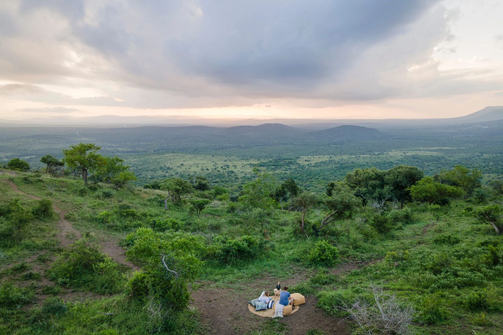 Two people recline on a blanket at a hilltop clearing above a wide green valley under a clouded dusk sky.