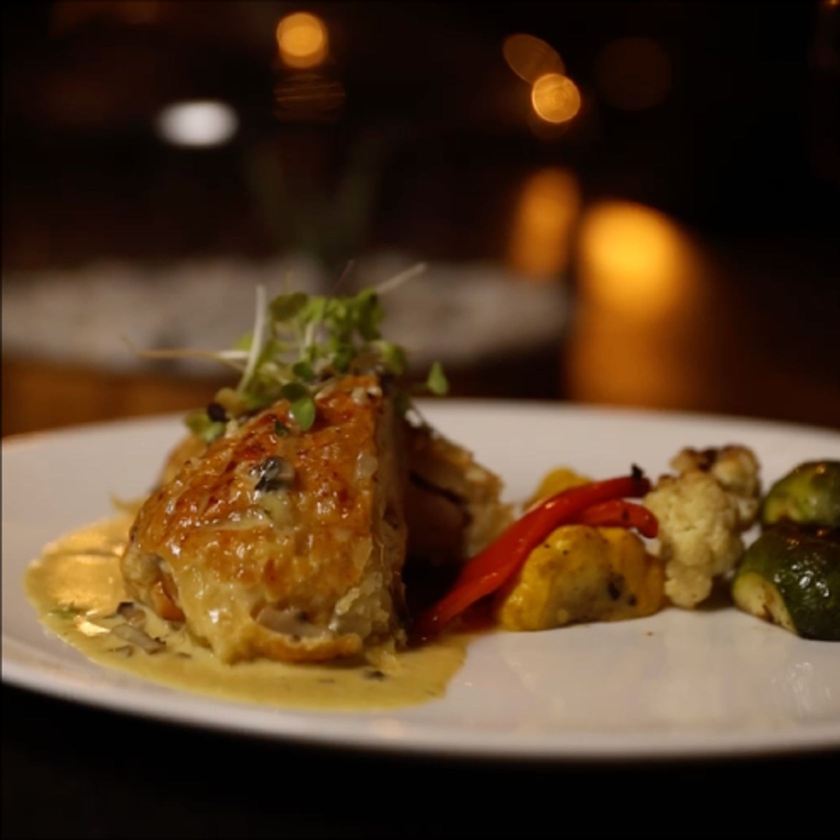 A seared chicken dish with roasted vegetables and cream sauce on a white plate by candlelight.