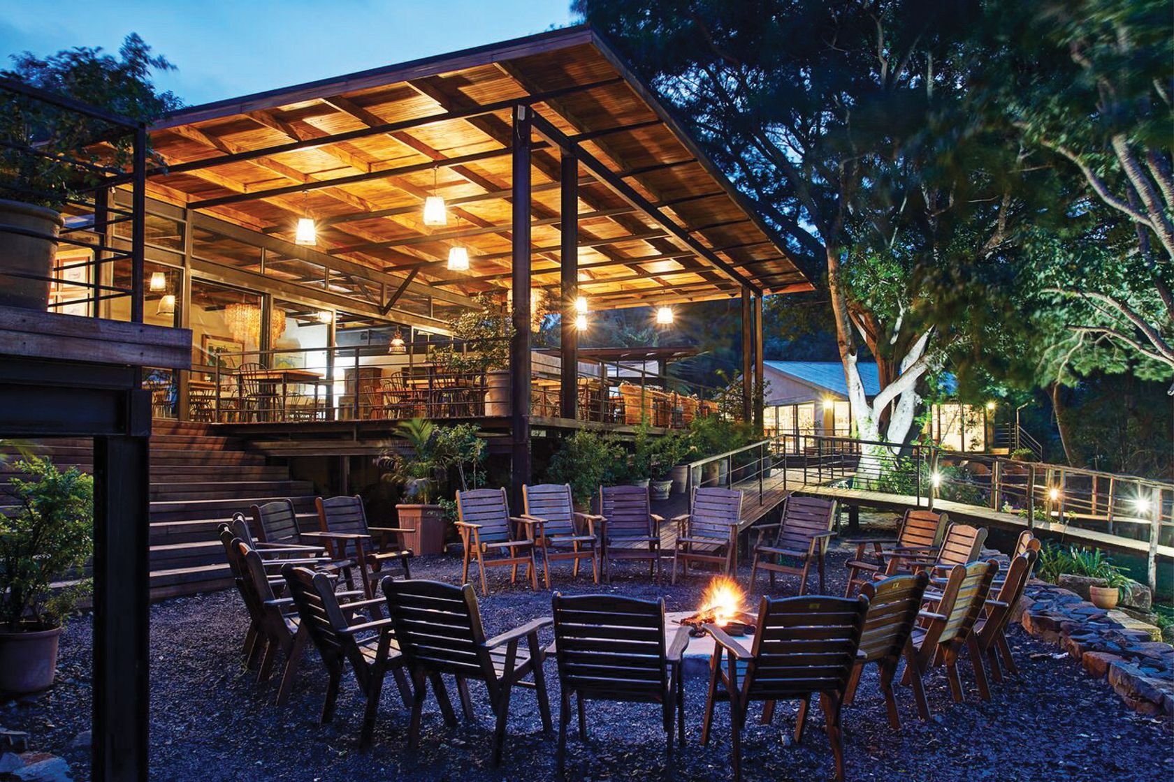A fire pit ringed by wooden chairs on gravel at dusk, with the lit timber lodge and a large fig tree behind.