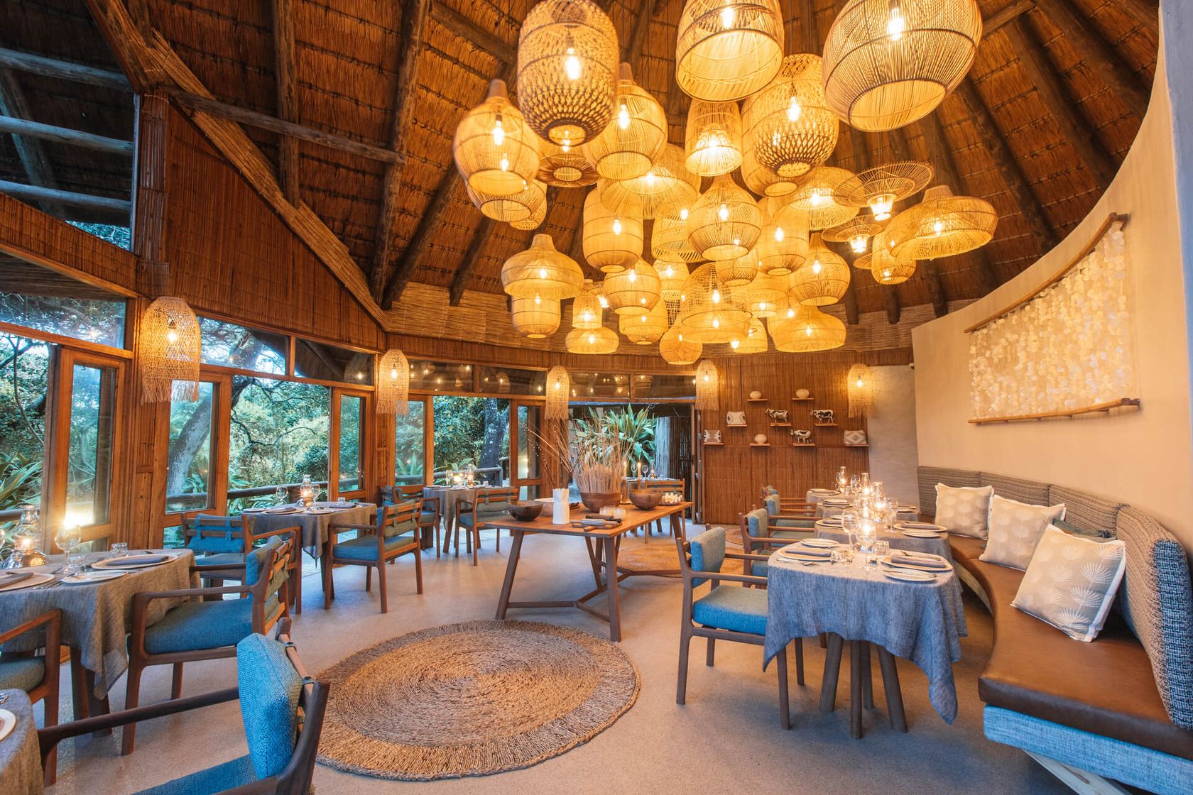 Restaurant interior at dusk under a thatched roof, a cluster of woven pendant lamps above blue-clothed tables and cushioned chairs.