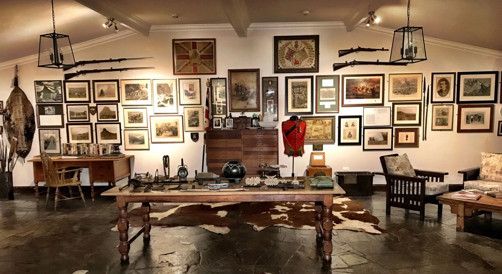 The lodge museum room, with framed battlefield prints, rifles mounted on the walls, a red military tunic, a Zulu shield, and a long wooden table over a cowhide rug.