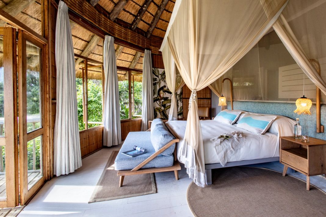 A four-poster bed under a mosquito net with blue pillows, reed-and-thatch walls and every window framing green forest.