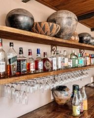 A bar shelf lined with spirit bottles and hanging wine glasses below African pottery and a woven basket.