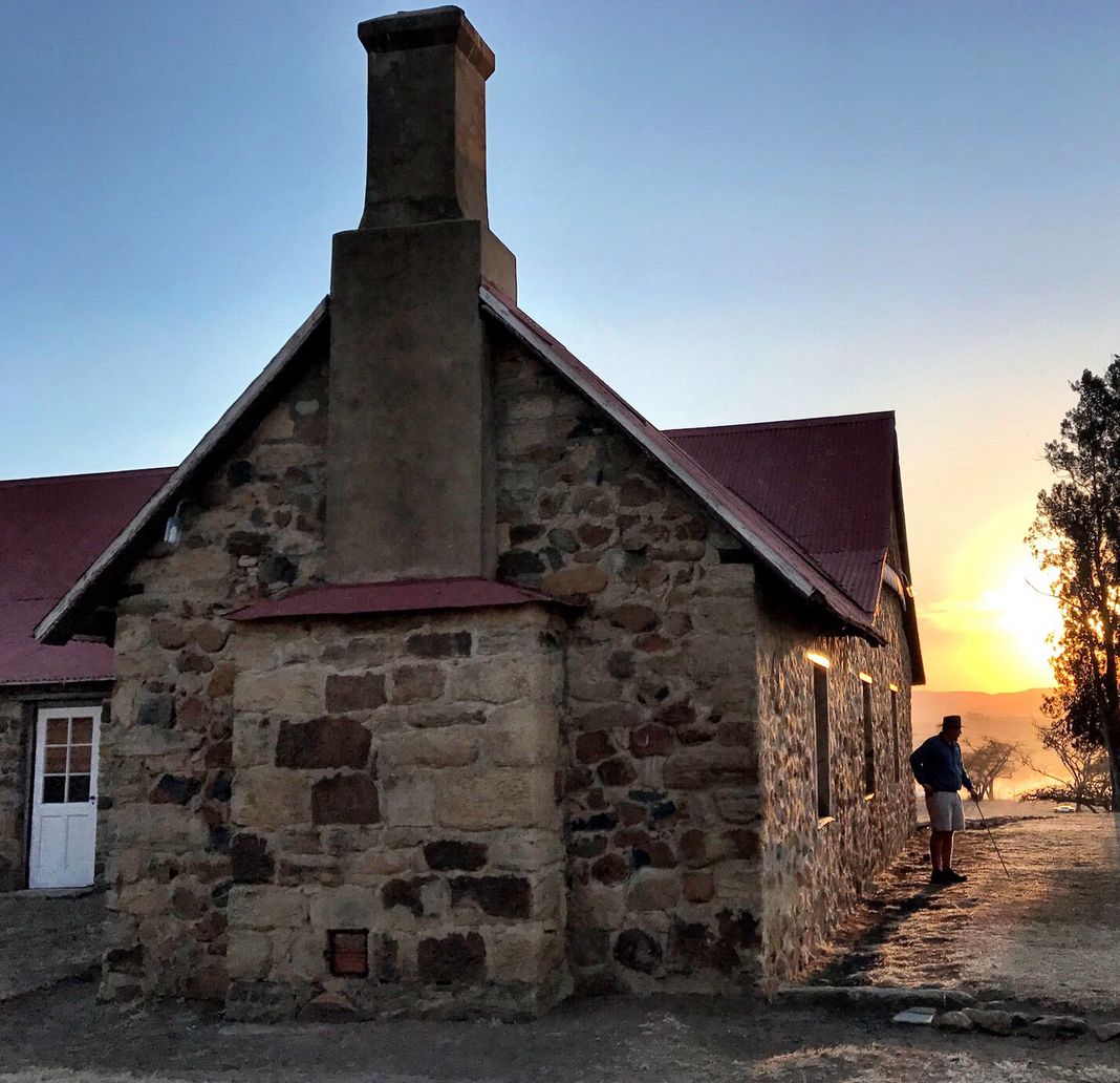 A lodge guide with a walking stick stands beside the old stone building with a red roof and tall chimney at Rorke’s Drift as the sun sets behind a tree.
