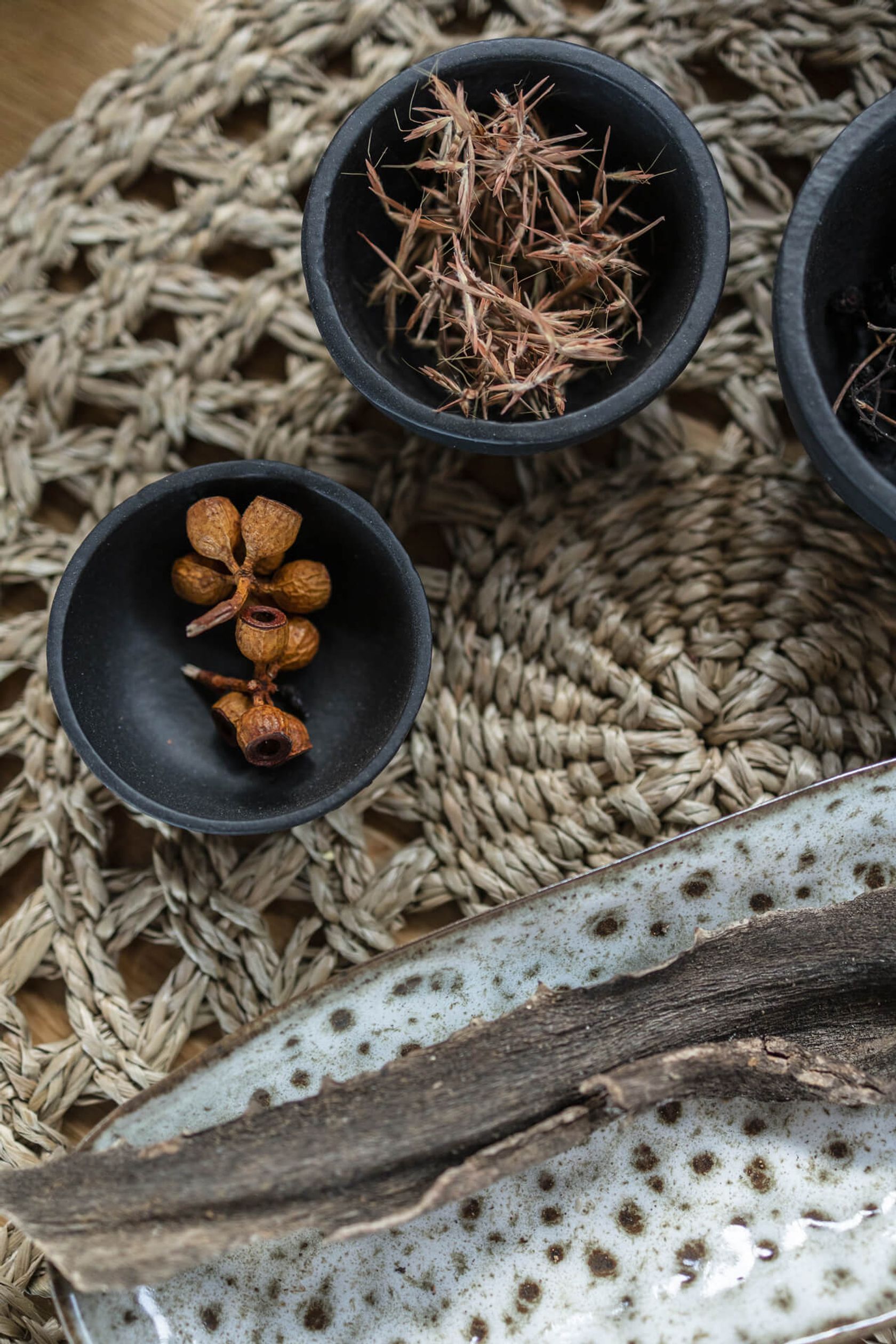 Dark bowls hold dried seed pods and grasses on a woven mat, seen from above.