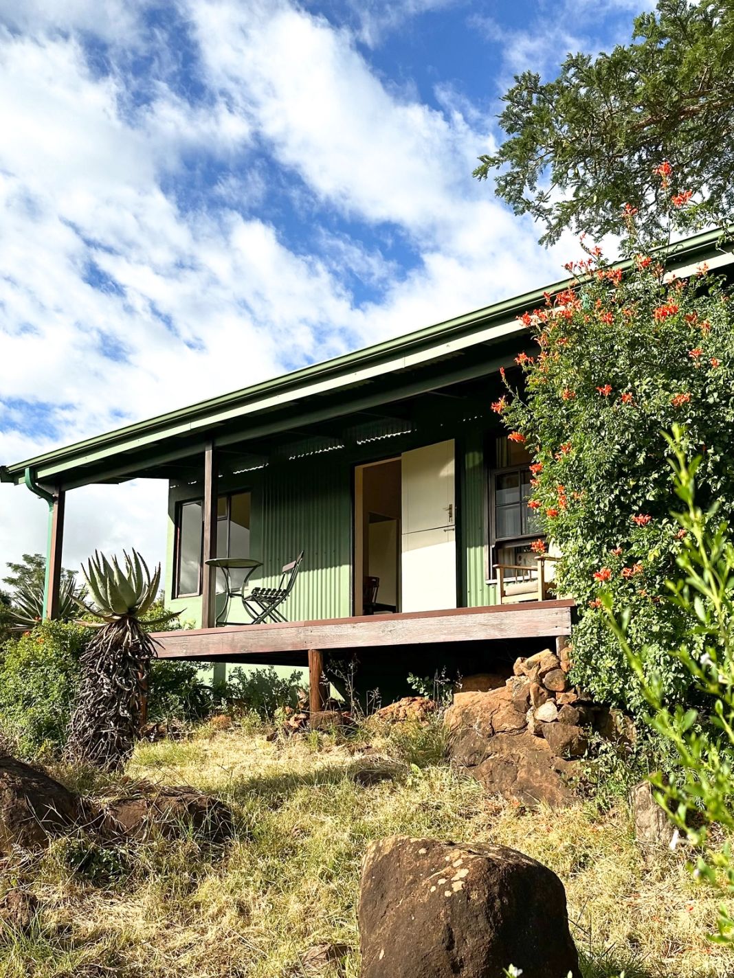 A green wood-and-iron cottage raised on a timber deck, with a bistro table on the veranda, aloes and a flowering orange shrub, under a partly clouded sky.