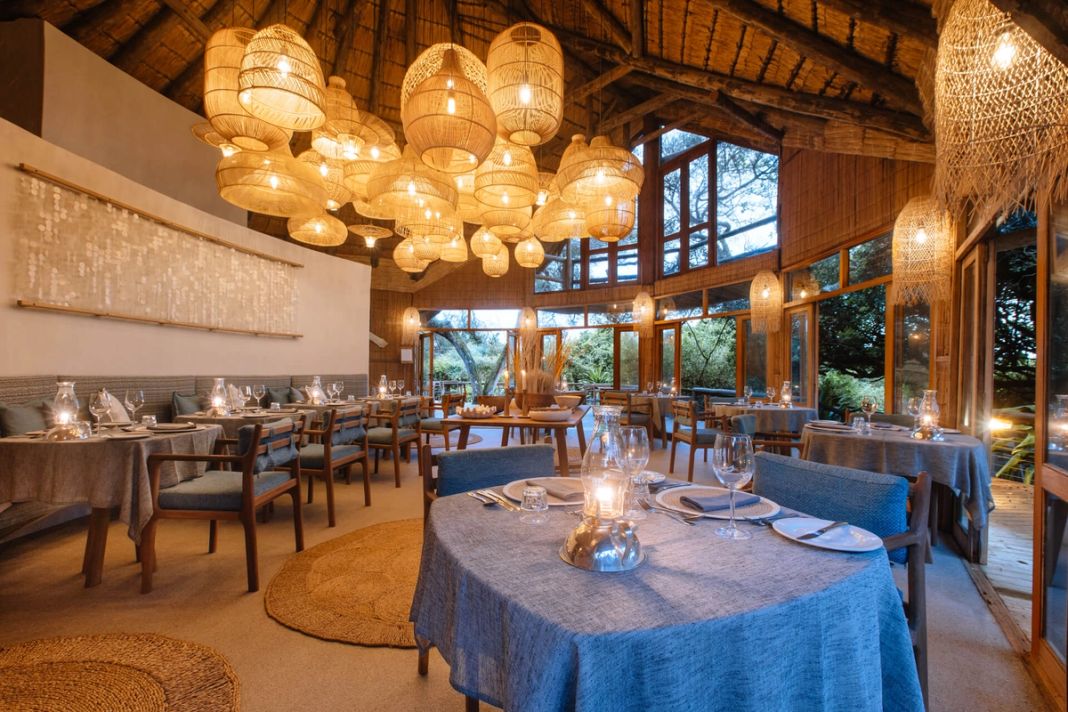Restaurant interior at dusk, blue-linen tables receding toward tall windows, woven pendant lamps and a shell-bead wall hanging.
