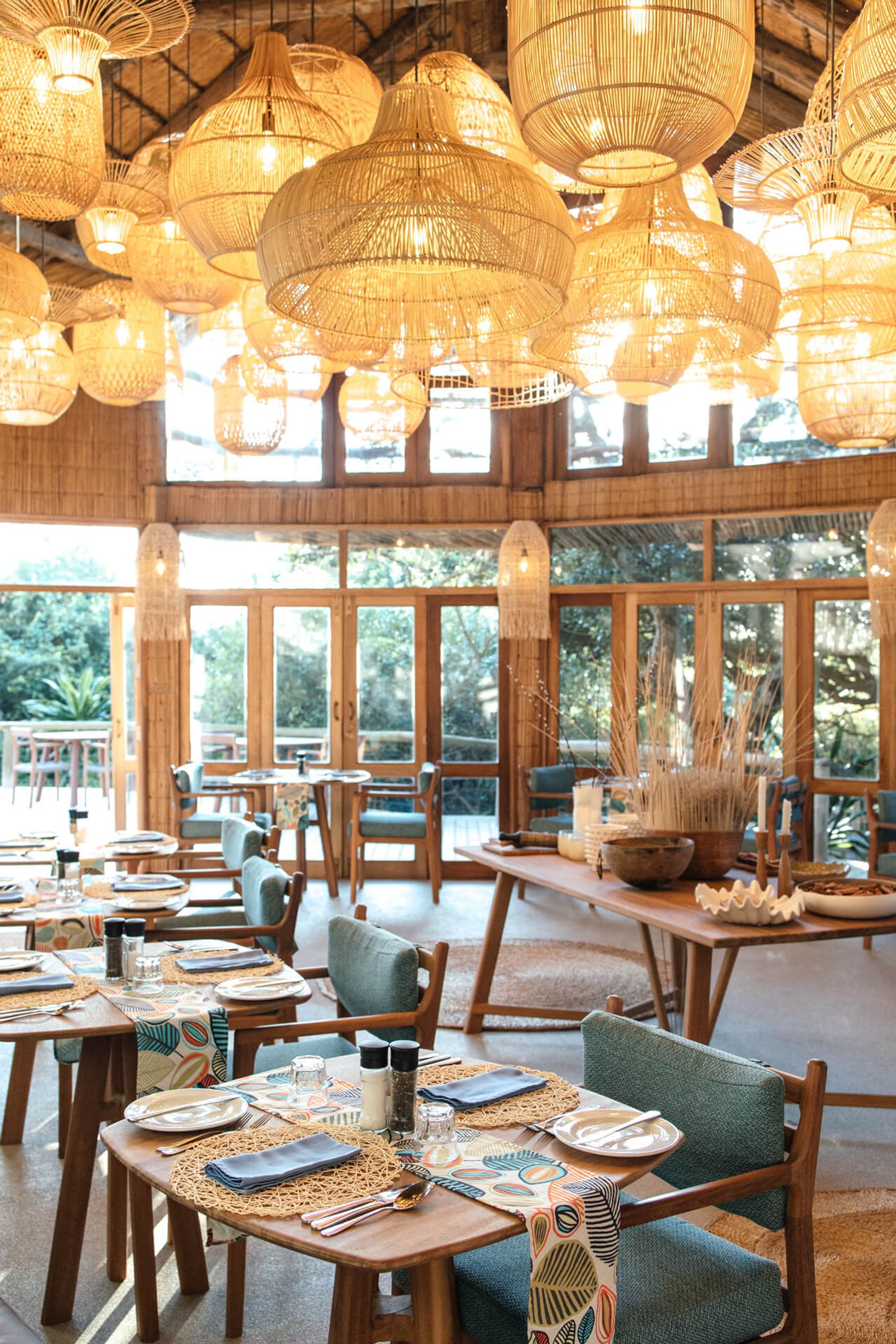 Restaurant interior by day, woven pendant lamps above wooden tables with cane placemats and teal chairs, windows to forest.