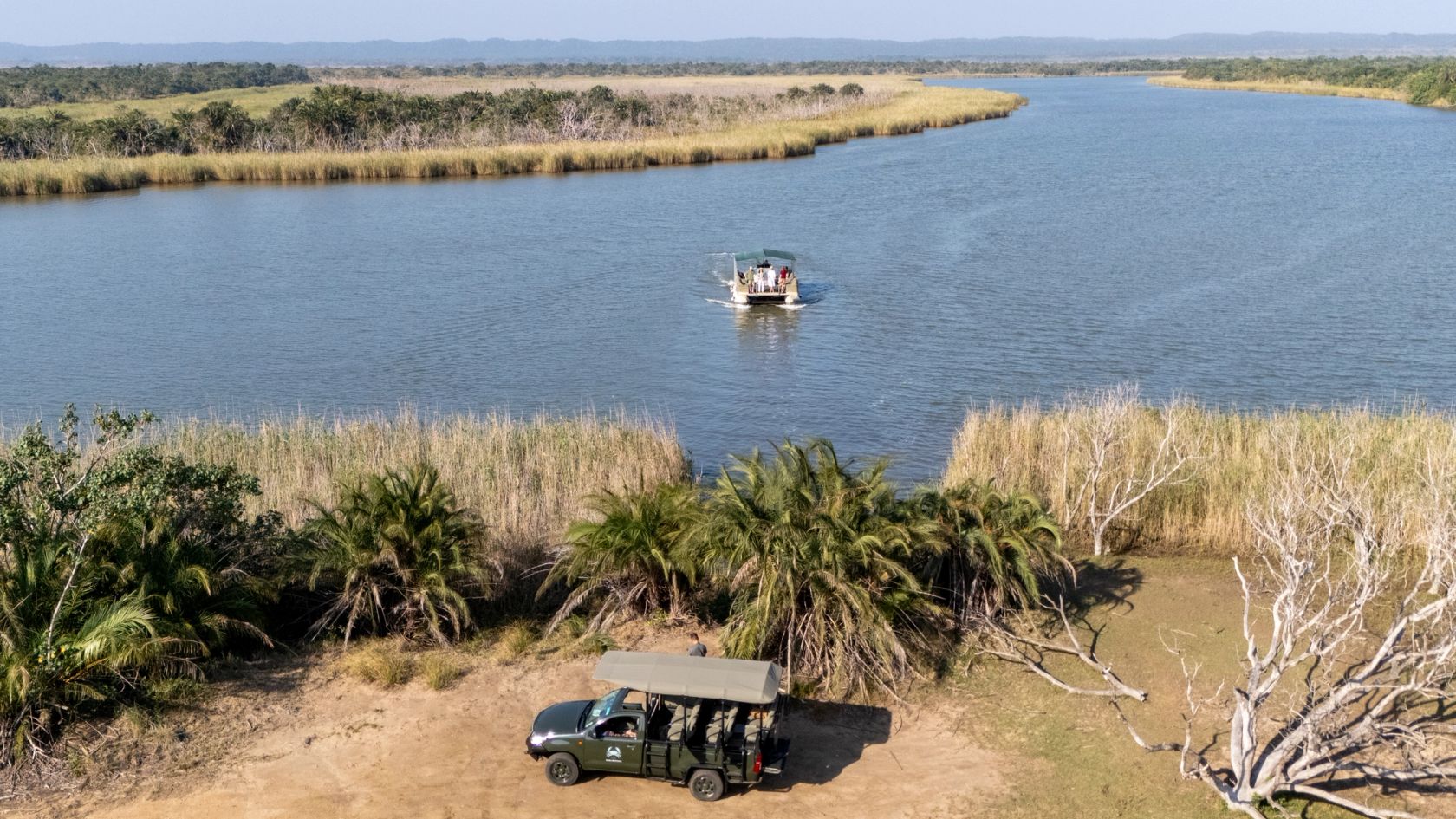 A solar-powered eco-boat crosses the estuary channel while a game-drive vehicle waits at the water's edge.