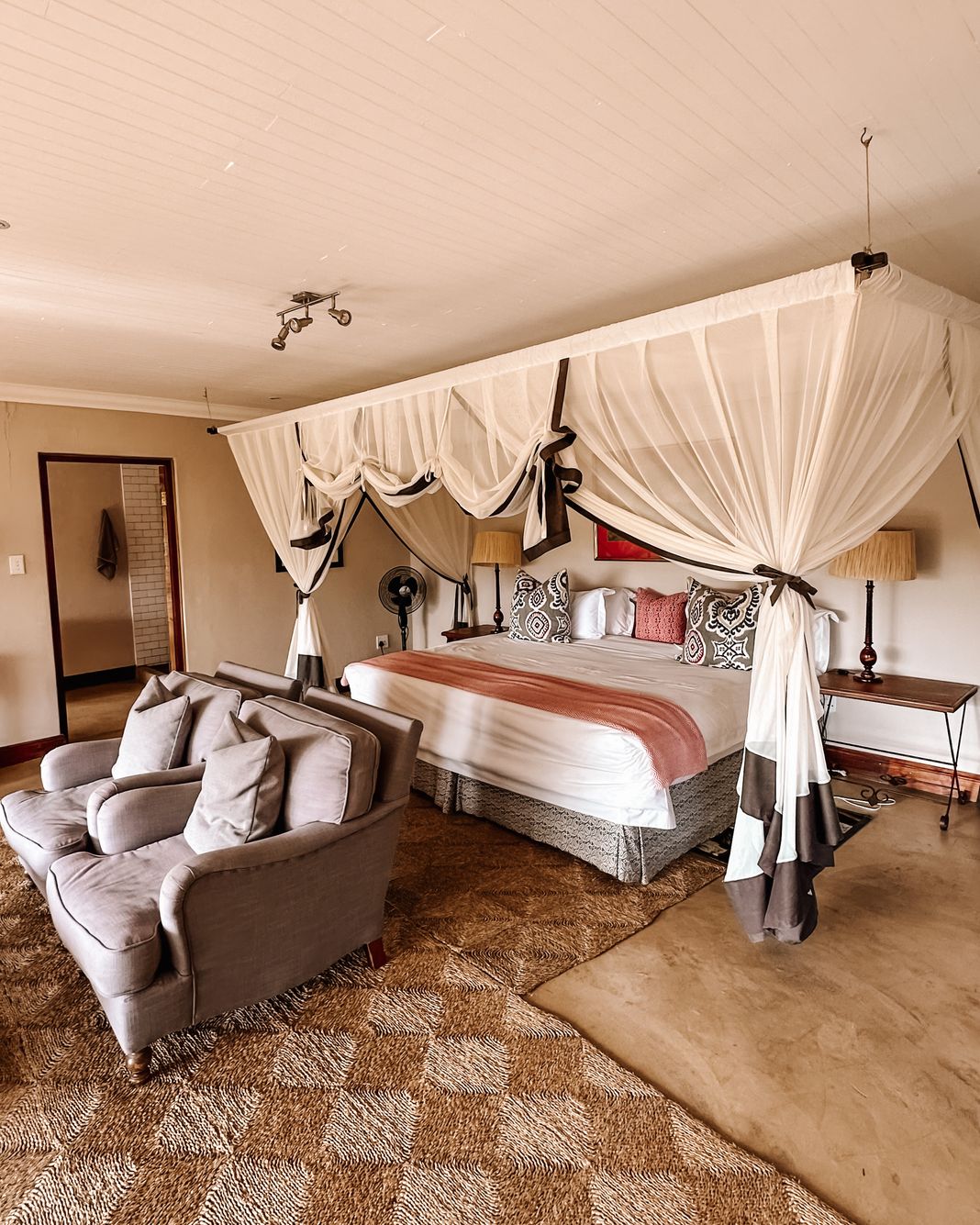 A bedroom with a four-poster bed draped in a mosquito net, a grey sofa and armchair, and a polished concrete floor, with an en-suite bathroom through the doorway.