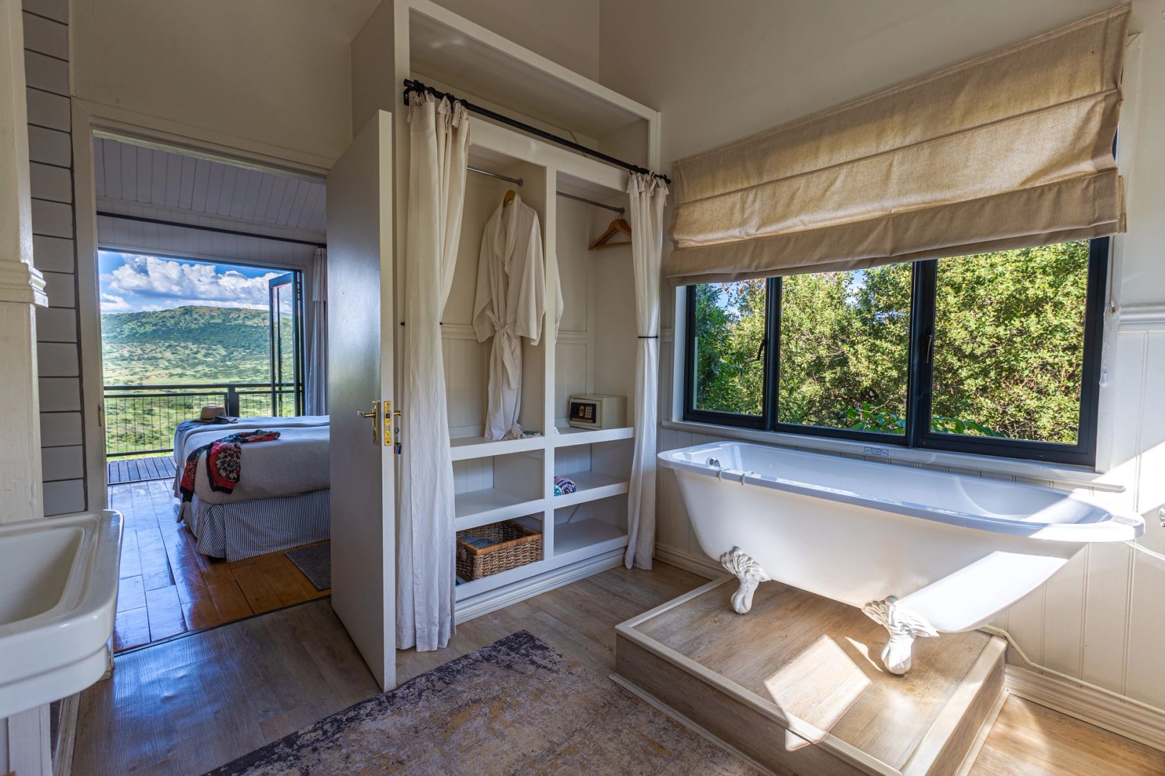 A clawfoot bath on a wooden step below a window of green foliage, with a robe in an open closet and a bedroom and valley view through the door.