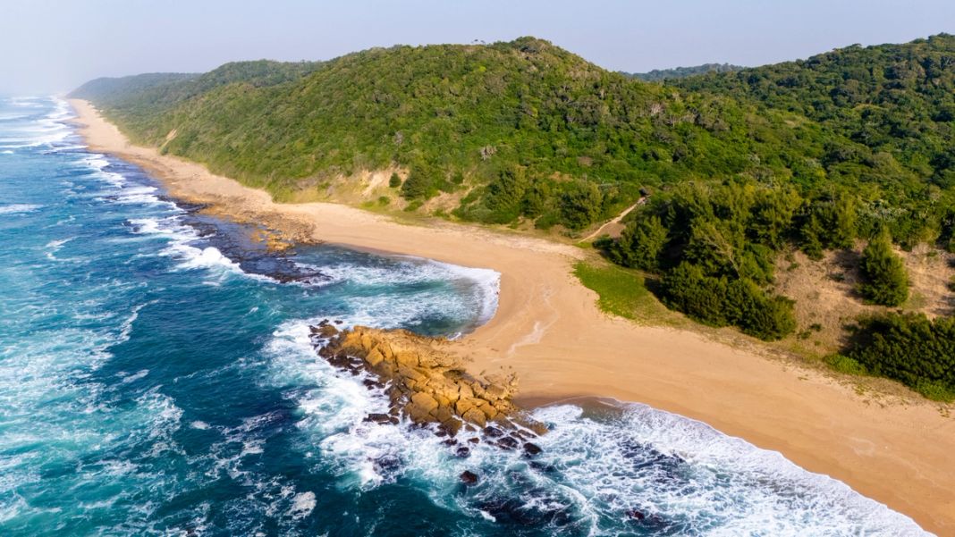 Aerial view of a deserted iSimangaliso beach where forested dunes meet a rocky point and the Indian Ocean surf.
