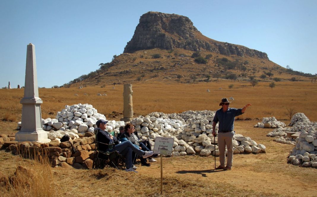 A guide in a hat points toward the Isandlwana battlefield while three visitors sit and listen beside a white memorial and whitewashed stone cairns, with the sphinx-shaped mountain behind.