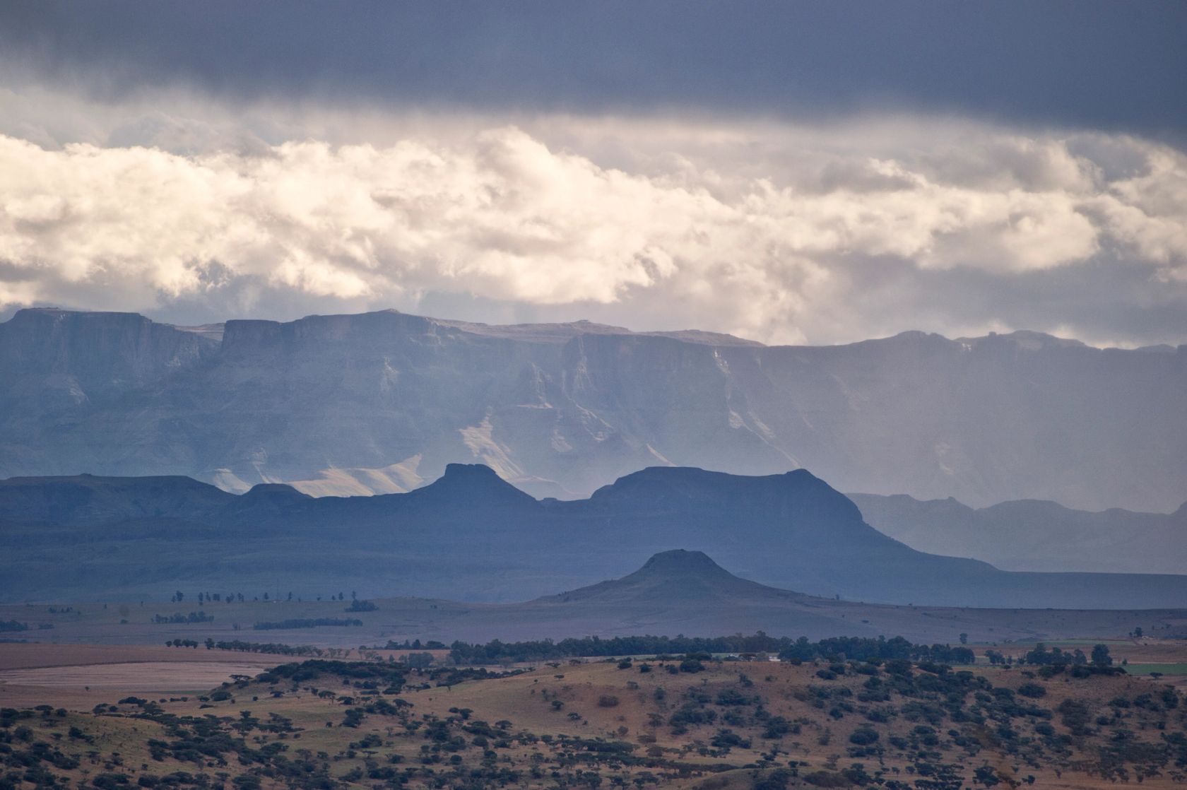 The drakensberg escarpment under storm cloud, layered blue ridges above a flat-topped koppie and tree-scattered veld.
