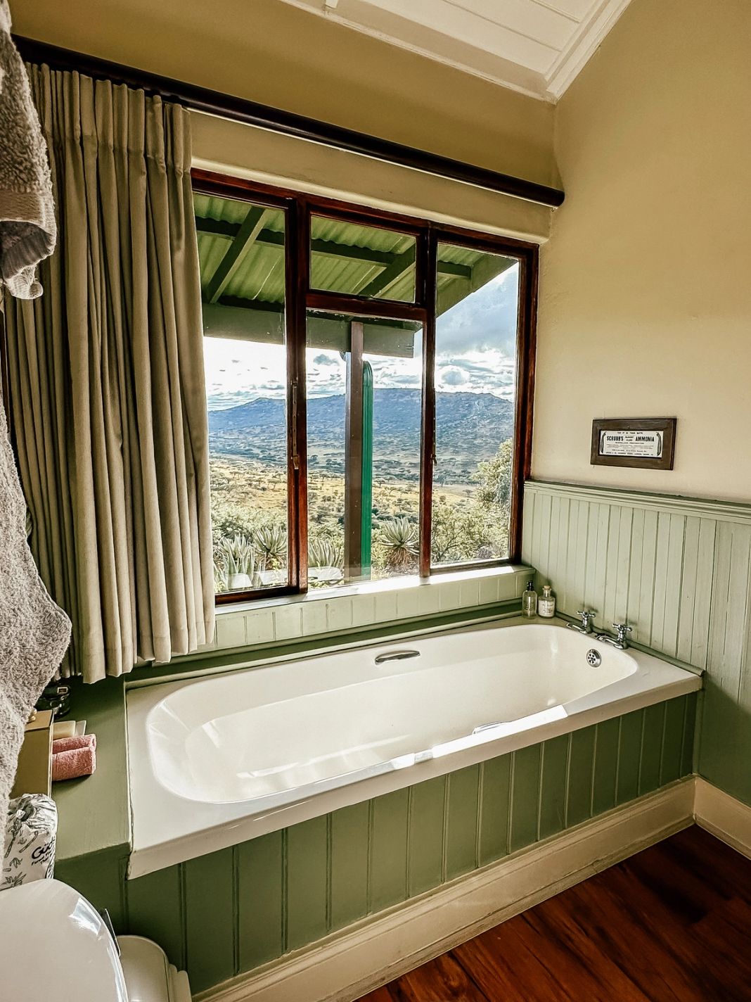 A cottage bathroom with a white built-in tub below a large window that frames aloes and a distant mountain range, set against green panelling.