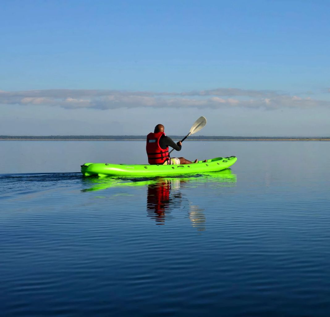 A lone paddler in a red life vest crosses the mirror-calm surface of Lake Sibaya in a green kayak.