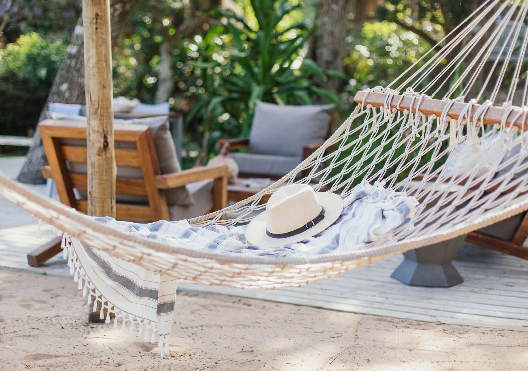 A white rope hammock draped with a striped throw and a straw hat, a cushioned wooden chair and greenery behind.