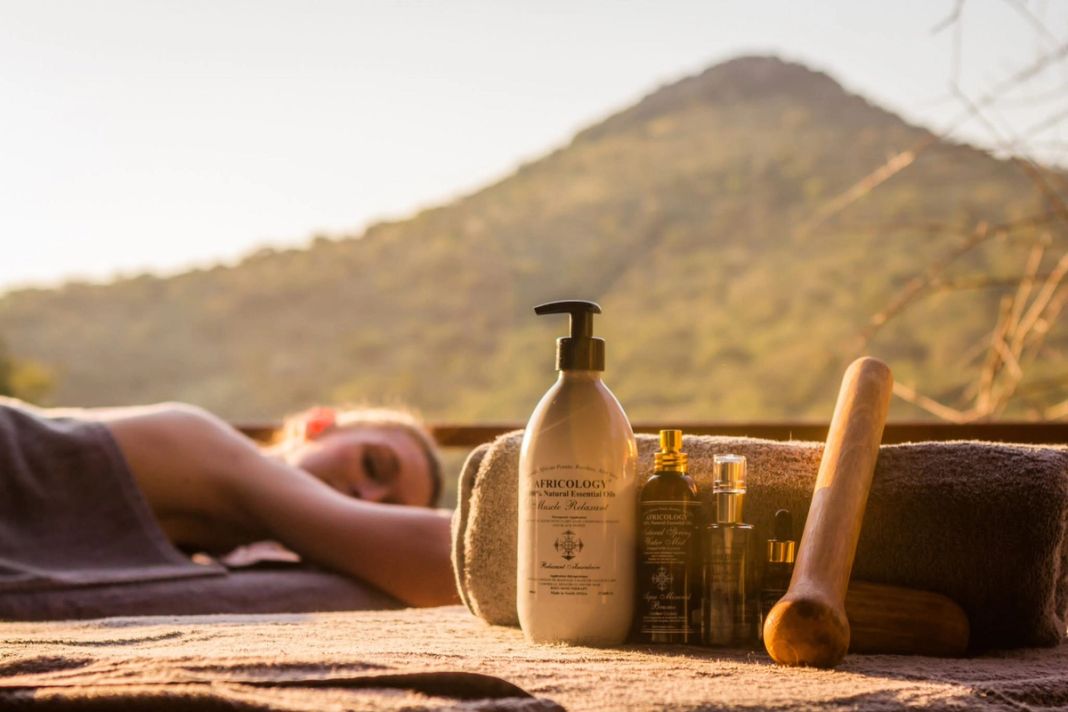 Africology massage oils rest on rolled towels beside a spa bed with a guest lying at rest and a hill beyond.