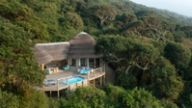 Golden-hour aerial of thatched huts spread across a densely forested slope, two small pools at the lower right.