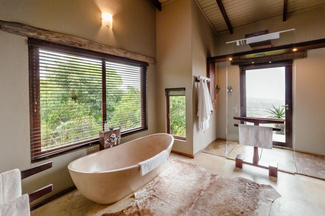 Villa bathroom with a freestanding stone tub, blind-covered windows, a cowhide rug and a bush view.
