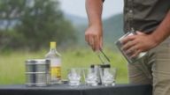 A ranger prepares gin and tonics with ice in the bush, glasses set out on a cloth-covered table.