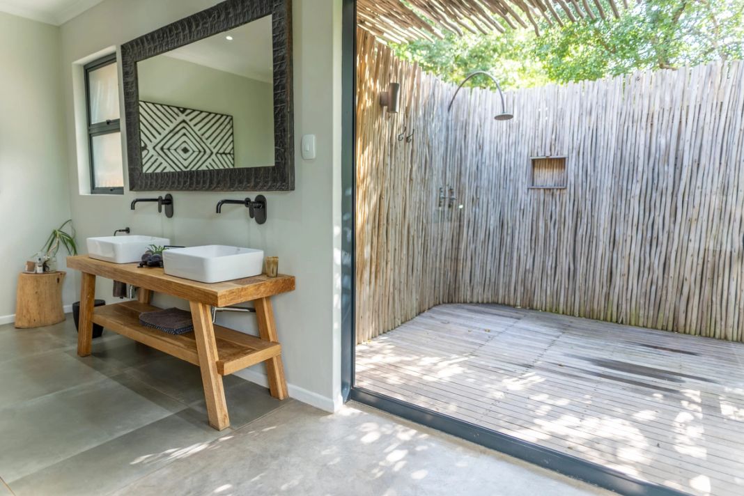 A suite bathroom with twin wall-mounted basins on a wood vanity opening through glass to a reed-walled outdoor shower.
