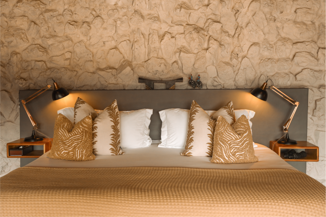 A suite bed dressed in cream and ochre against a rough stone wall with reading lamps.