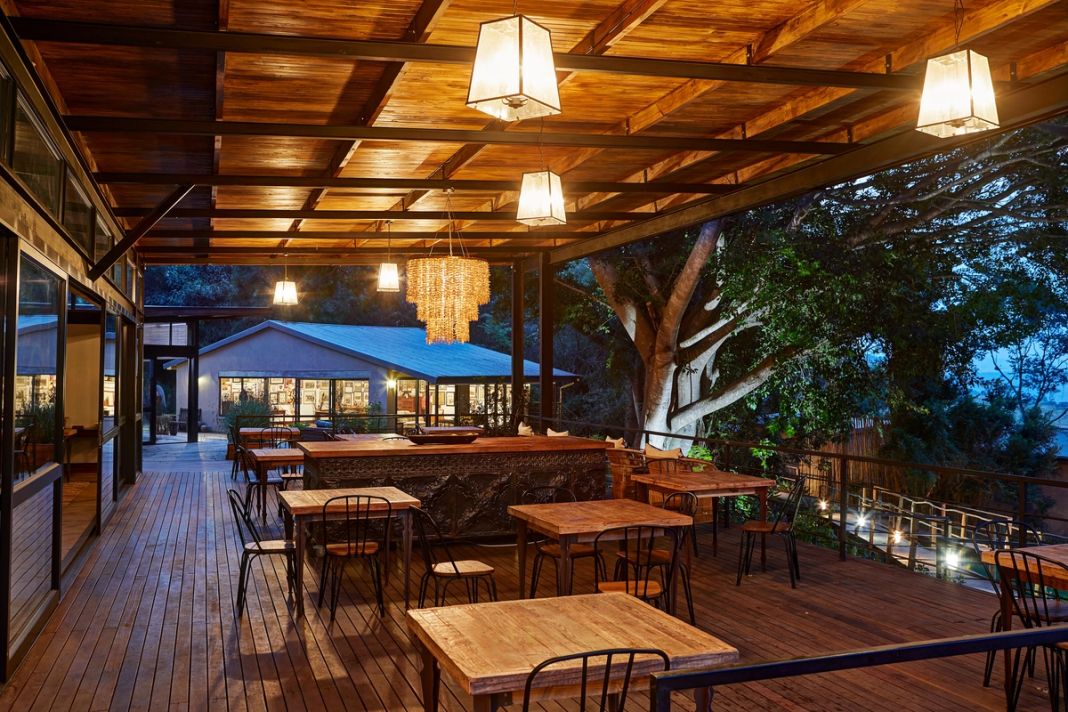 A covered timber dining deck at dusk with pendant lanterns, wooden tables and chairs, and a fig tree beside the lodge building.