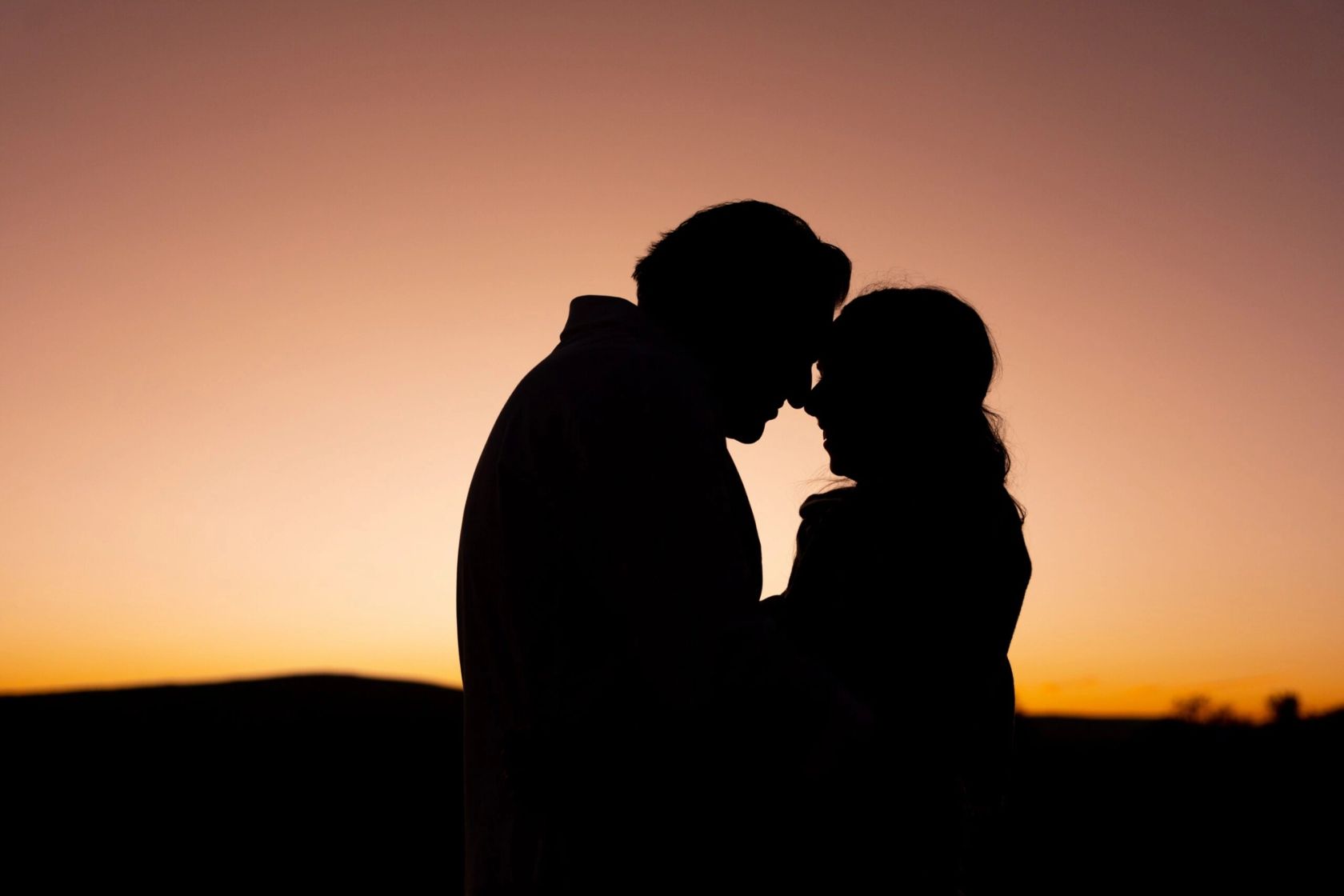 Two people stand forehead to forehead as silhouettes against an orange and pink sunset sky.