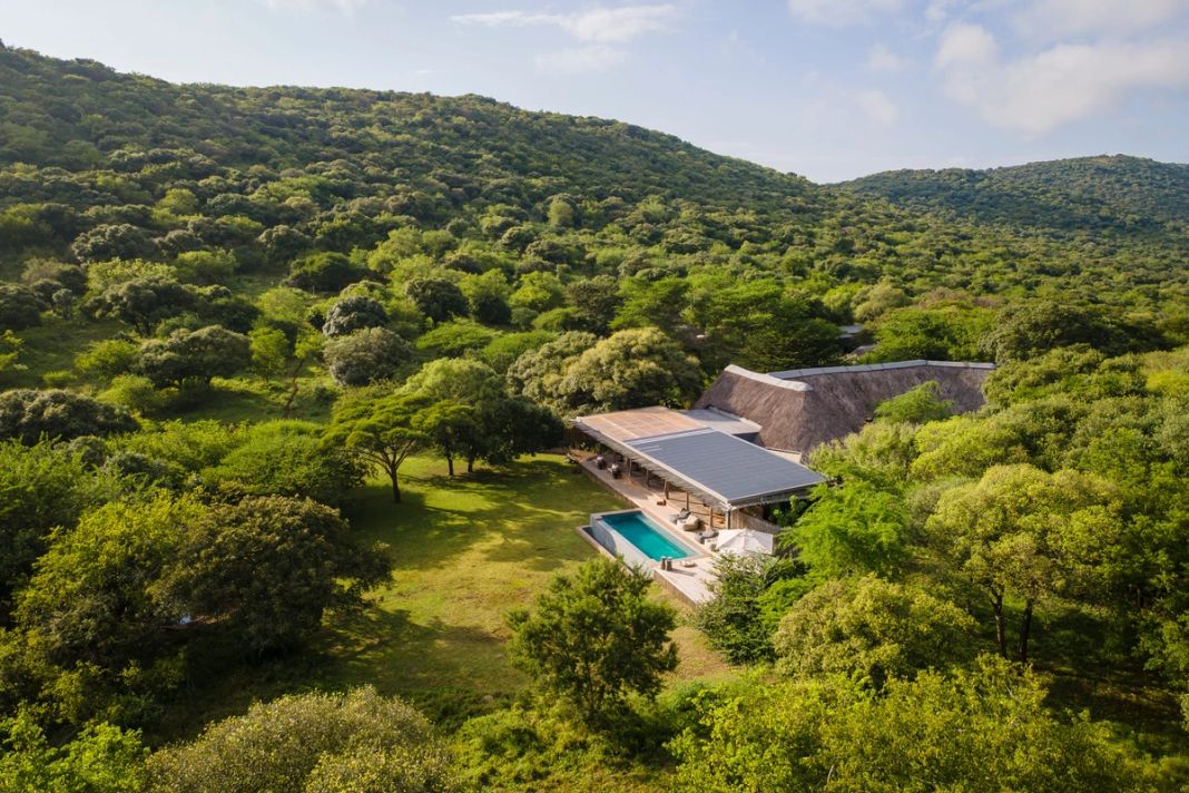 Aerial view of a thatched lodge and pool set in a clearing surrounded by green hills and scattered trees.