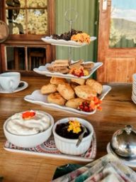 A three-tier stand of scones, biscuits and oat clusters with edible flowers, beside bowls of cream and jam on a wooden table.