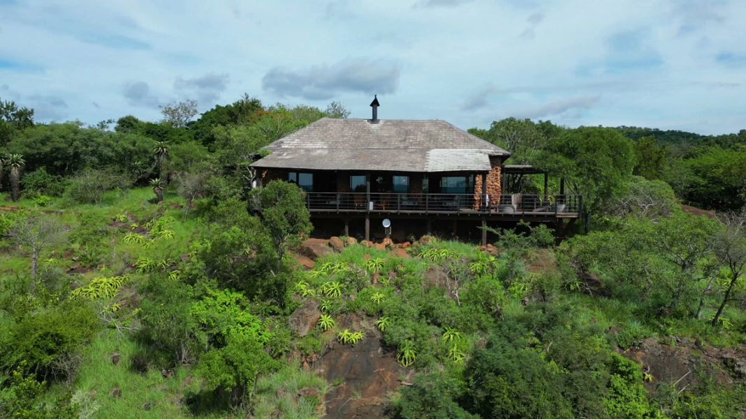 The stone-and-thatch main lodge stands on its clifftop among green bushveld under a blue sky.