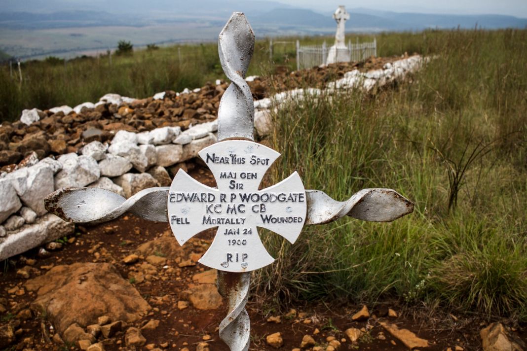 A white iron cross marks where Major General Woodgate fell in 1900, with a stone-walled grave and a Celtic cross on the grass ridge behind.