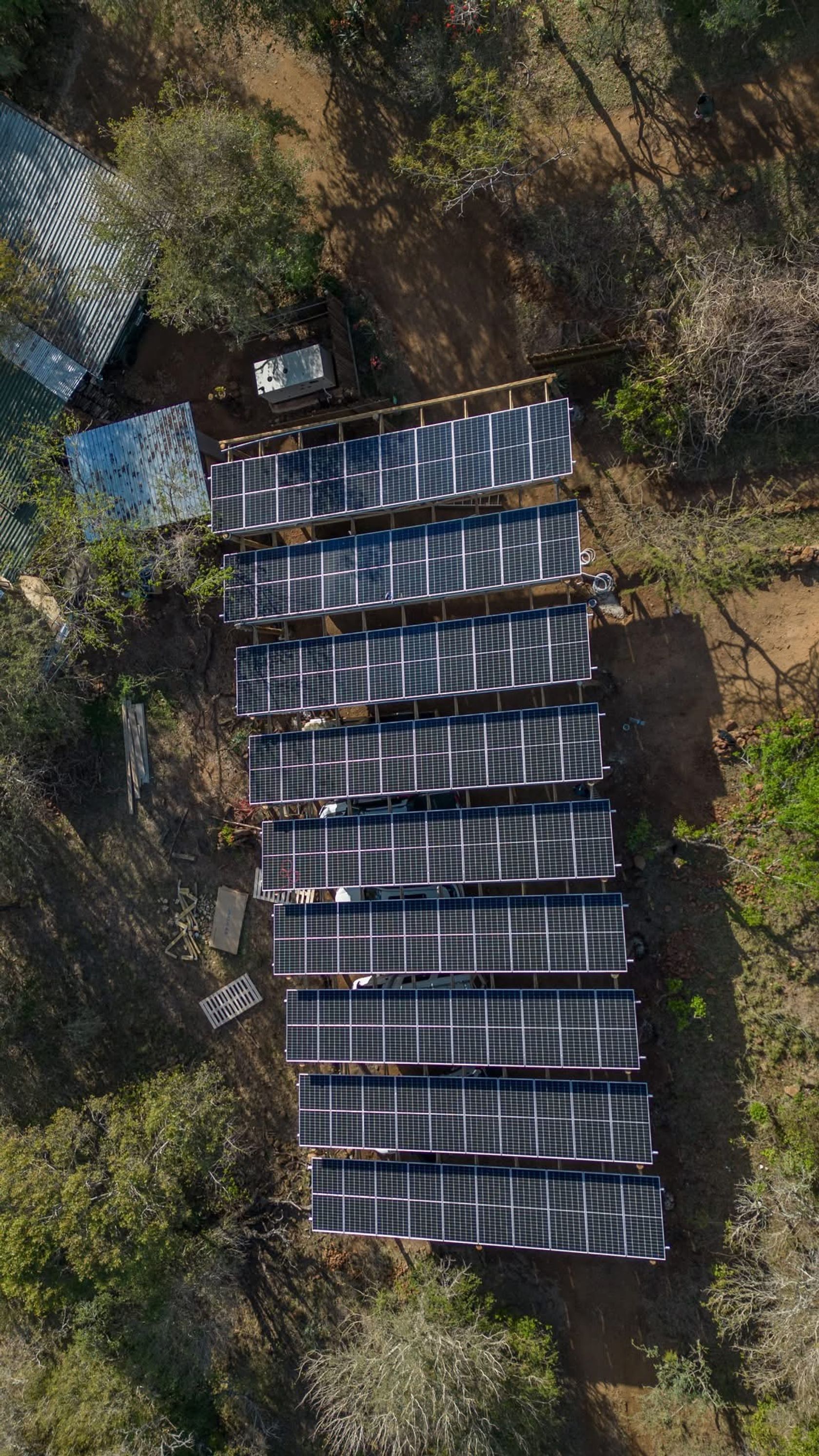 Overhead aerial view of rows of solar panels set among bushveld trees and dirt tracks.