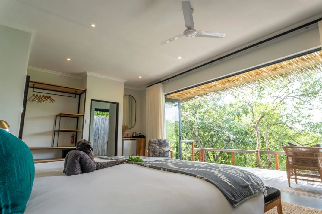 A king bed with a ceiling fan and open shelving faces glass stack-back doors that open onto a forest deck.