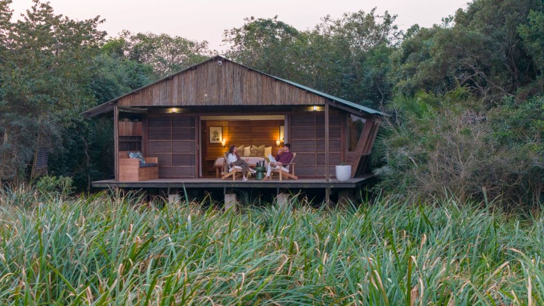 Two guests sit on the lit veranda of the honeymoon suite at dusk, the cabin standing alone in tall wetland reeds.