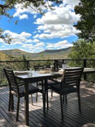 A dining table set with wine glasses and plates on a wooden deck, overlooking green Drakensberg foothills under a cloud-scattered sky.