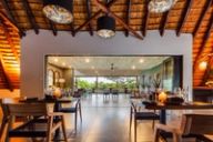 Set dining tables with candles sit under a thatched roof, opening through glass doors to a veranda and bushland view.