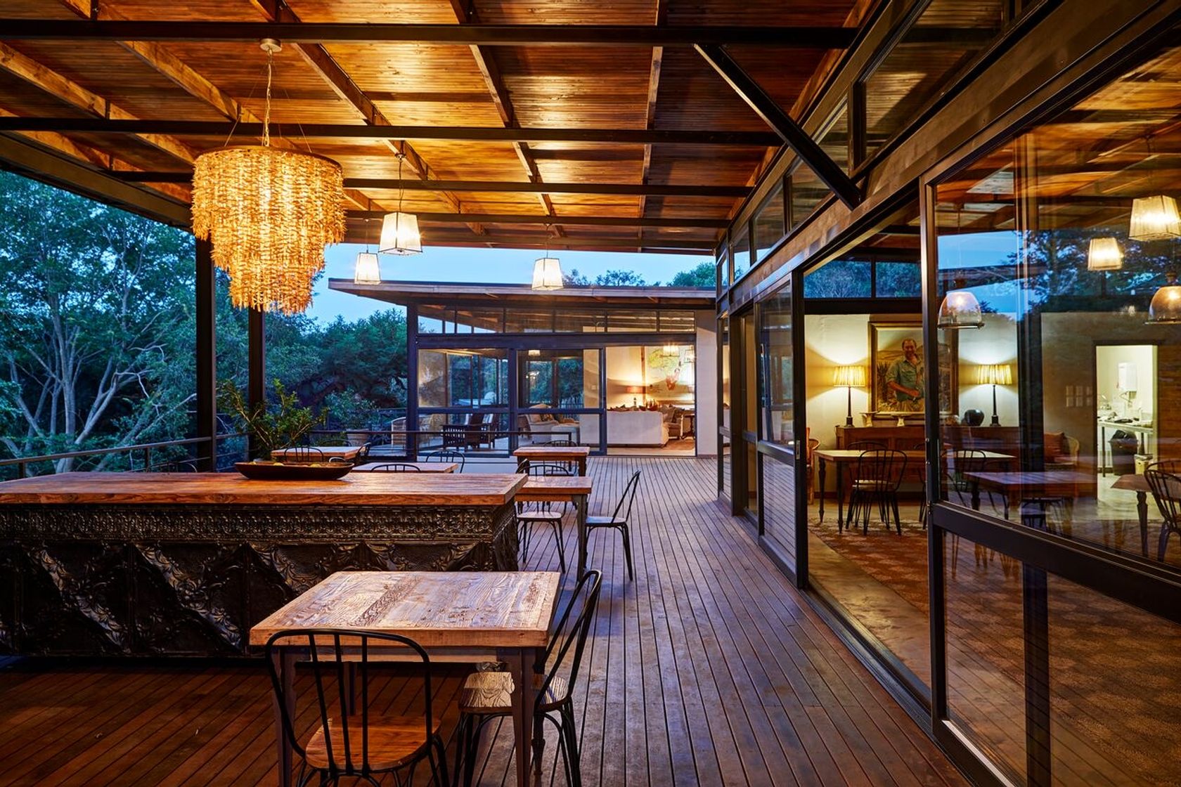 An open-sided timber dining deck at dusk with a beaded chandelier, high tables and metal chairs, and a lit interior lounge visible through glass doors.